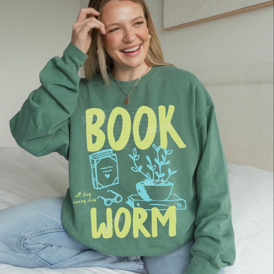 Person wearing a green sweatshirt with 'BOOK WORM' text and graphics, sitting on a bed.