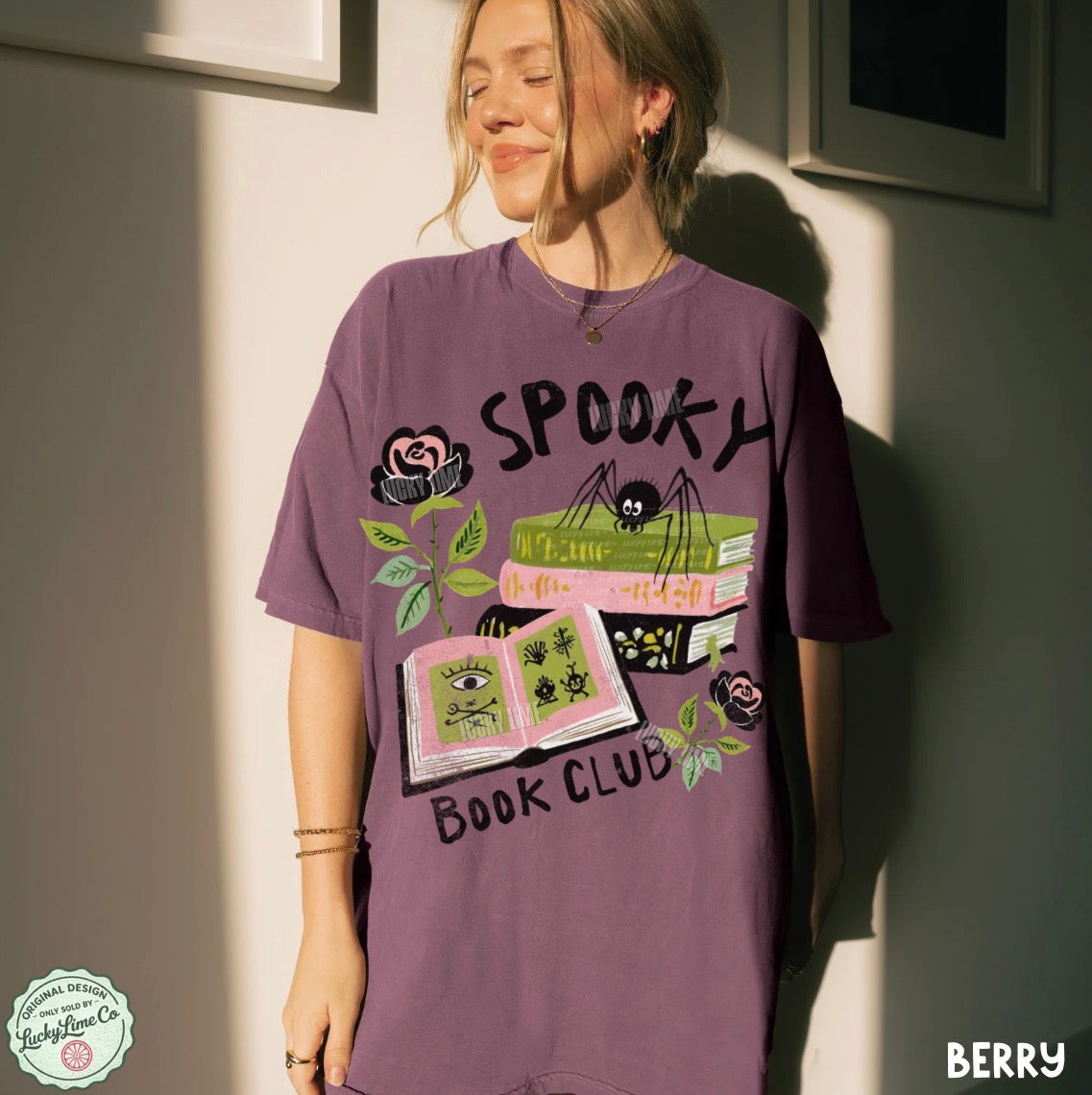Person wearing a purple 'Spooky Book Club' t-shirt against a neutral wall.
