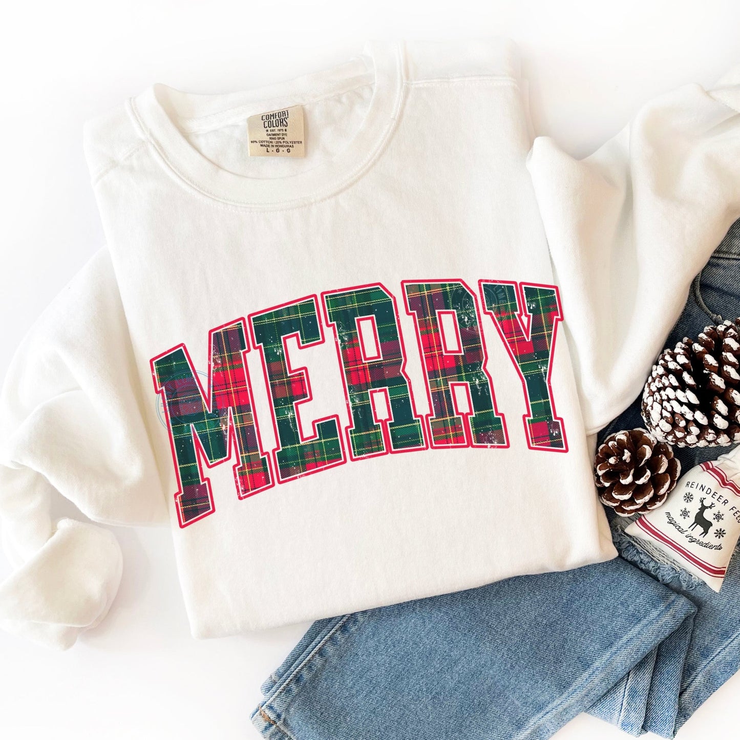 White sweatshirt with plaid 'MERRY' text, paired with jeans and pinecones on a white background.
