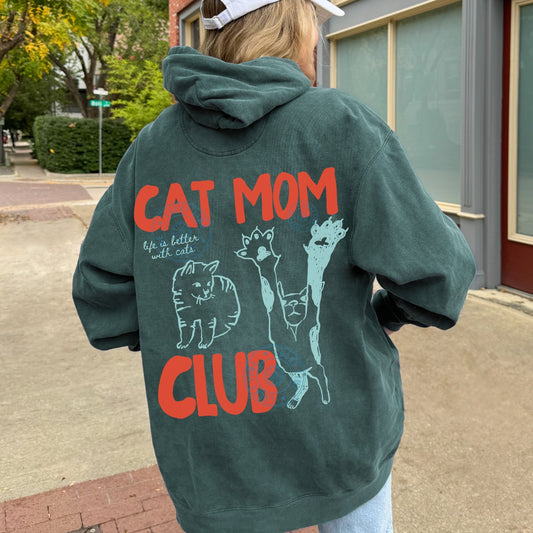 Person wearing a green hoodie with 'Cat Mom Club' text and graphics on the back, standing outdoors.