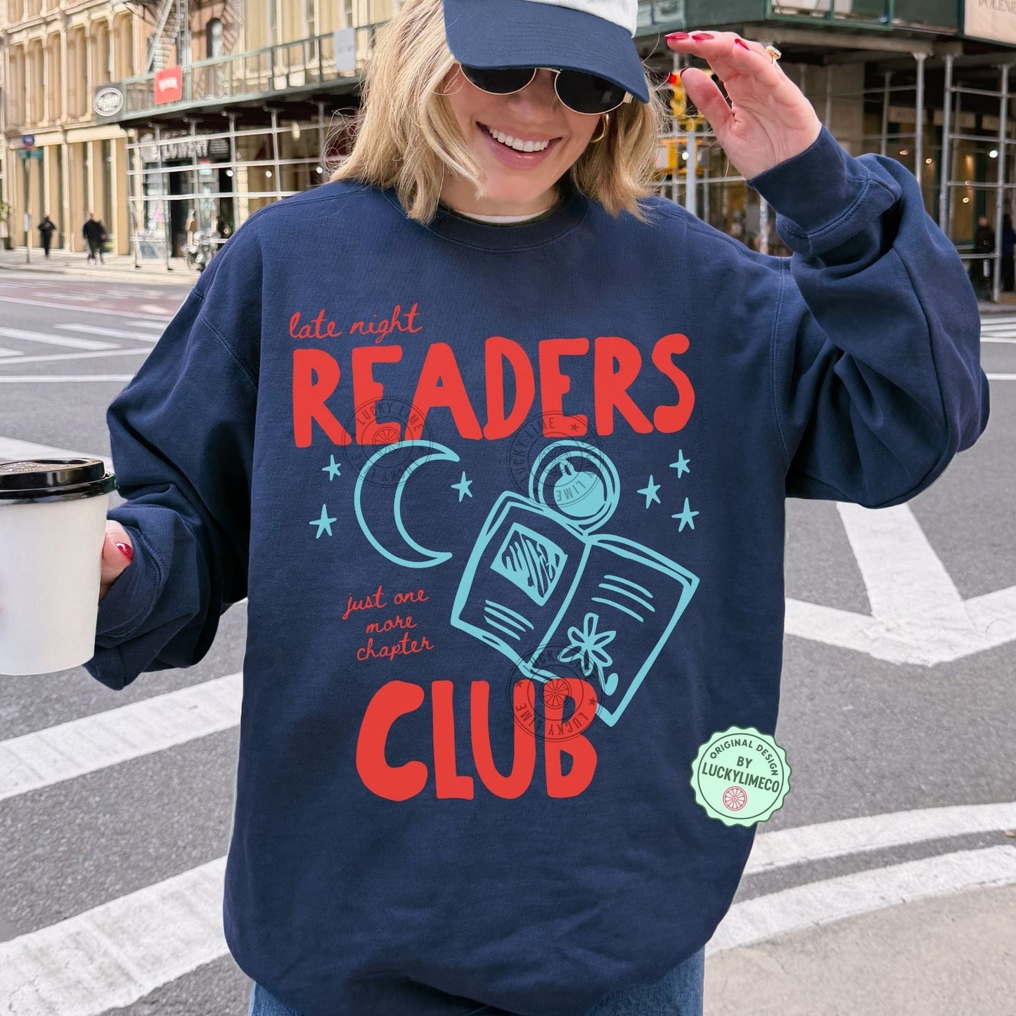 Person wearing a navy sweatshirt with 'Late Night Readers Club' design, holding a coffee cup on a city street.