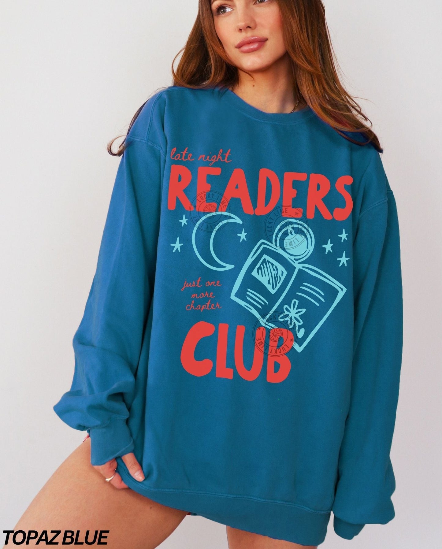 Person wearing a topaz blue sweatshirt with 'late night readers club' text and graphics on a plain background