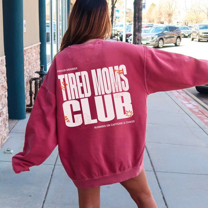Person wearing a maroon sweatshirt with 'Tired Moms Club' text on the back, standing on a sidewalk.