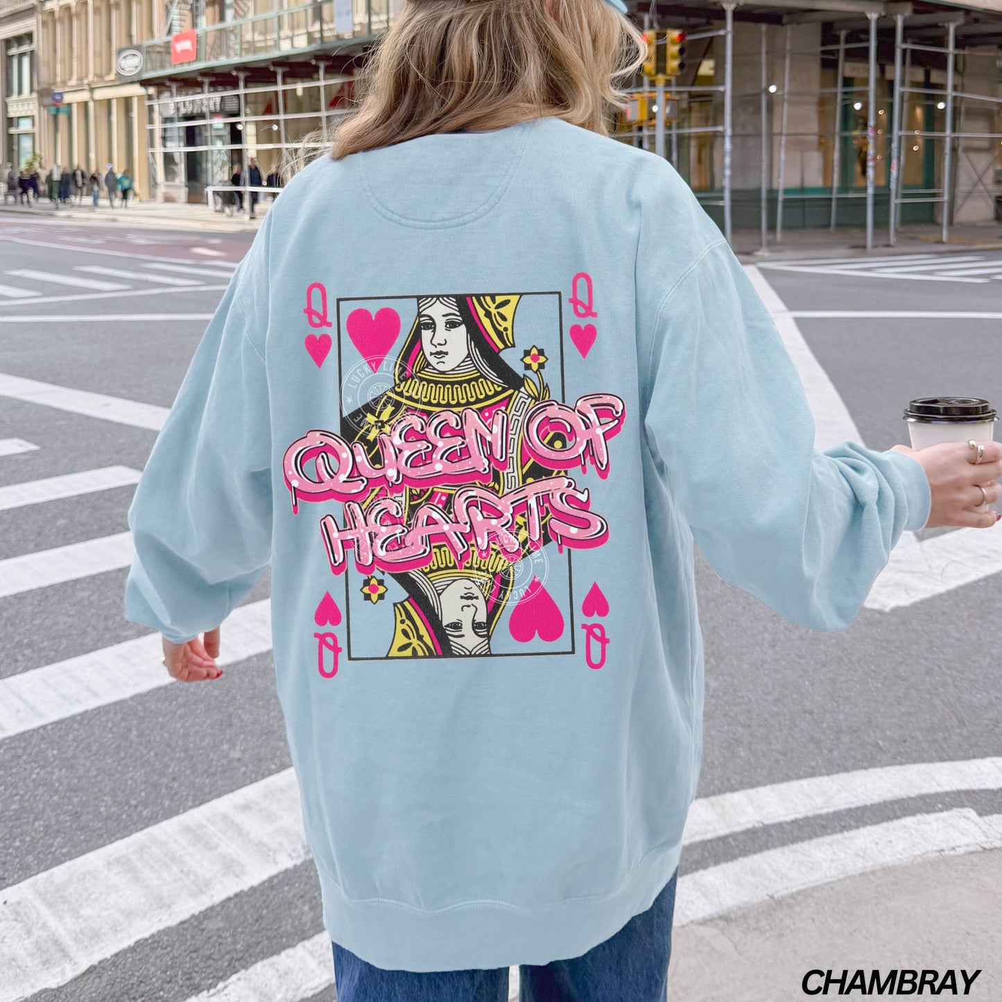 Person wearing a light blue sweatshirt with 'Queen of Hearts' design on a city street.
