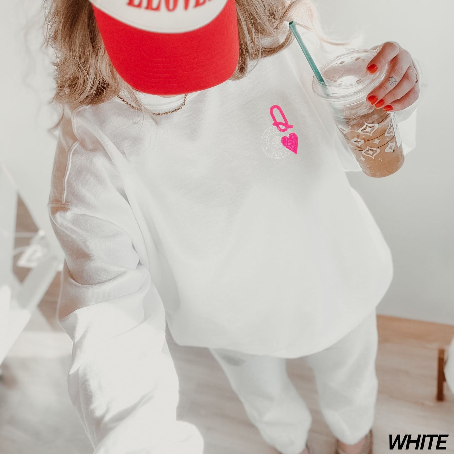 Person wearing a white sweatshirt with a pink logo, holding a clear cup with a straw.