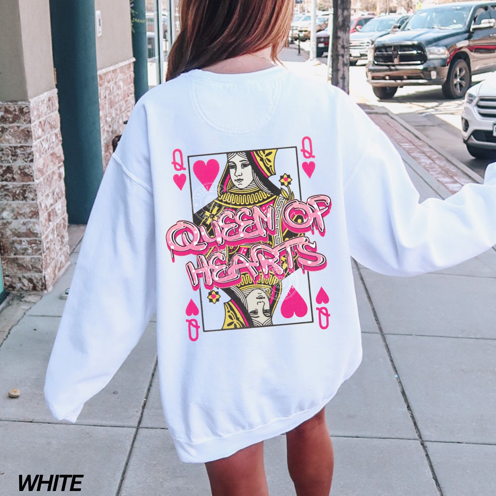 Person wearing a white sweatshirt with 'Queen of Hearts' design on the back, standing on a sidewalk.