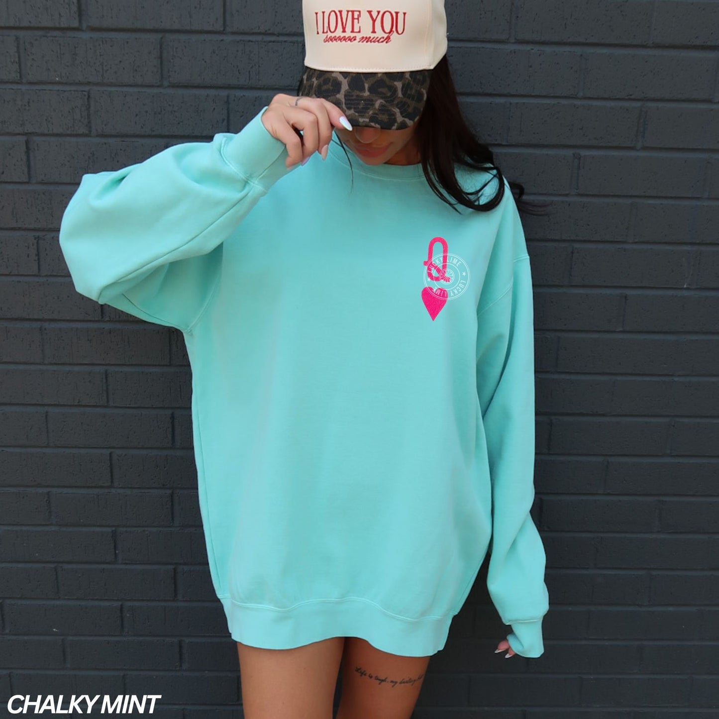 Person wearing a chalky mint green sweatshirt with a pink logo against a black brick wall.