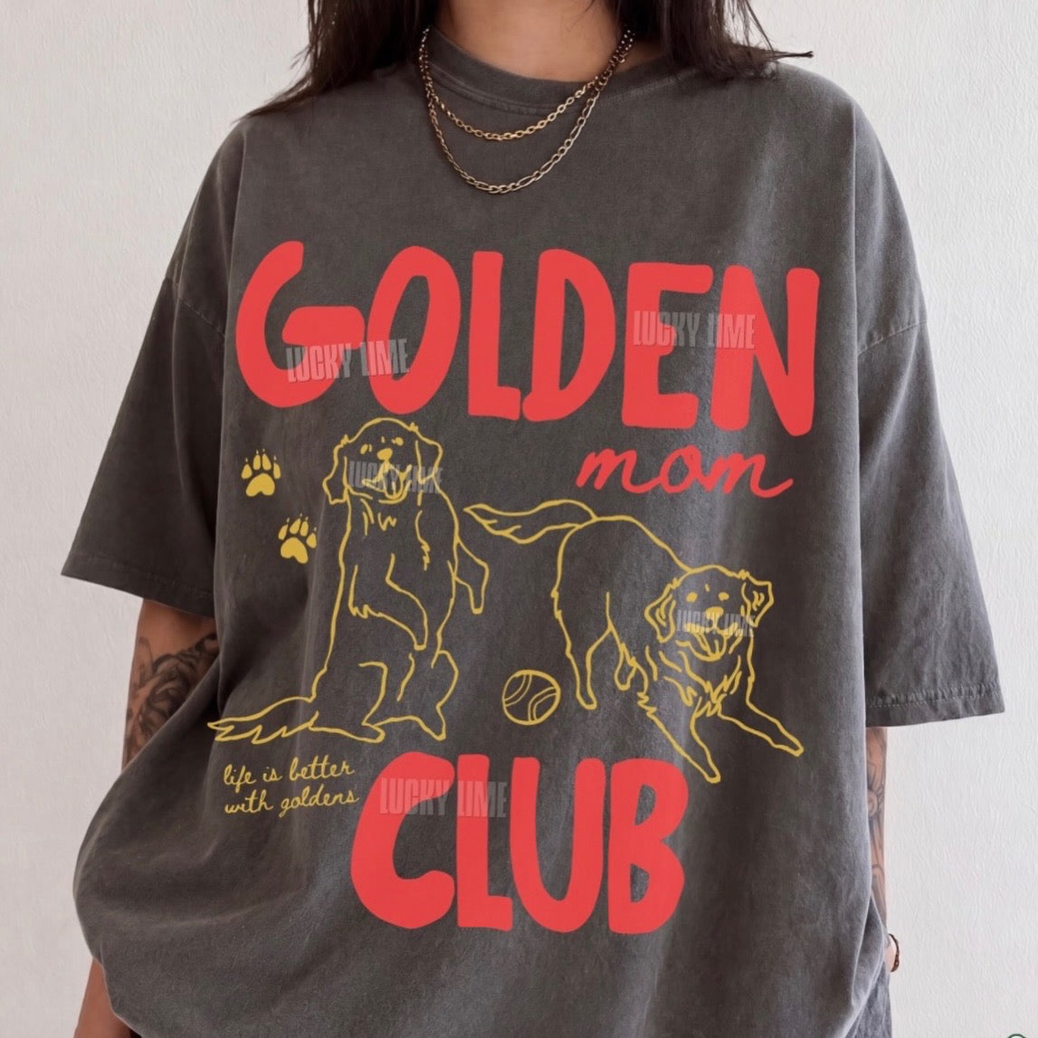 A person wearing an oversized dark gray graphic T-shirt with red and yellow text reading 'GOLDEN mom CLUB' and line drawings of two golden retrievers, photographed against a plain light wall.