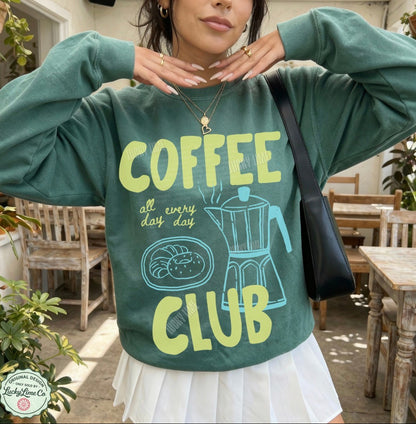 Person wearing a green sweatshirt with 'Coffee Club' text and design, standing outdoors.