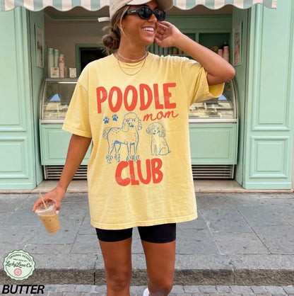 Woman wearing a yellow t-shirt with 'Poodle Mom Club' text and graphics, standing in front of a green building.