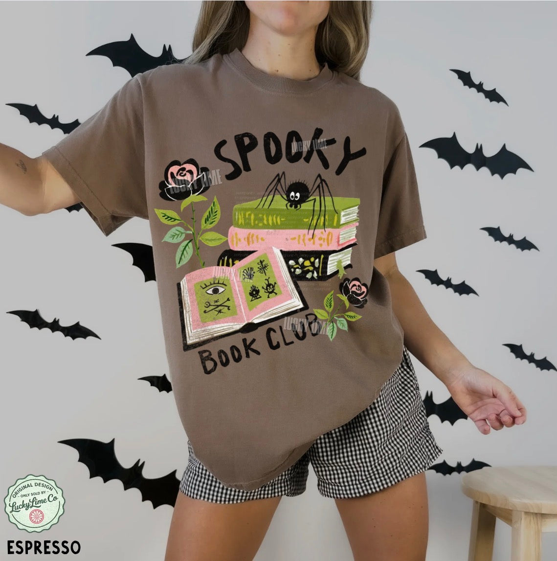 Person wearing a brown t-shirt with 'Spooky Book Club' design and checkered shorts, surrounded by bats.