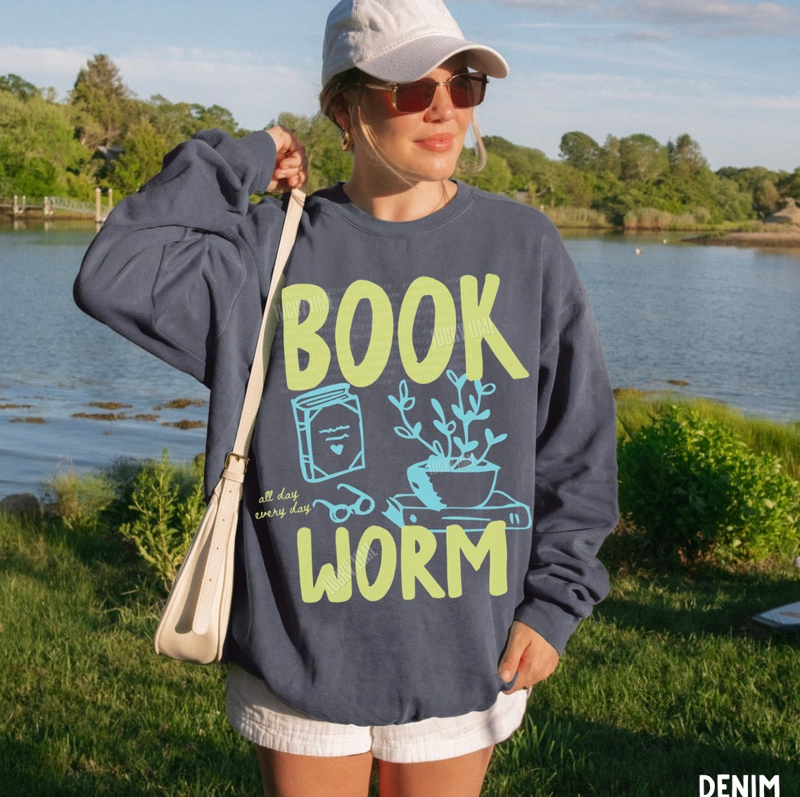 Person wearing a denim colored sweatshirt with 'BOOK WORM' text by a lake