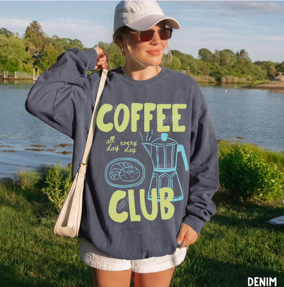 Person wearing a denim sweatshirt with 'Coffee Club' text by a lake
