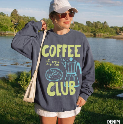 Person wearing a denim sweatshirt with 'Coffee Club' text by a lake