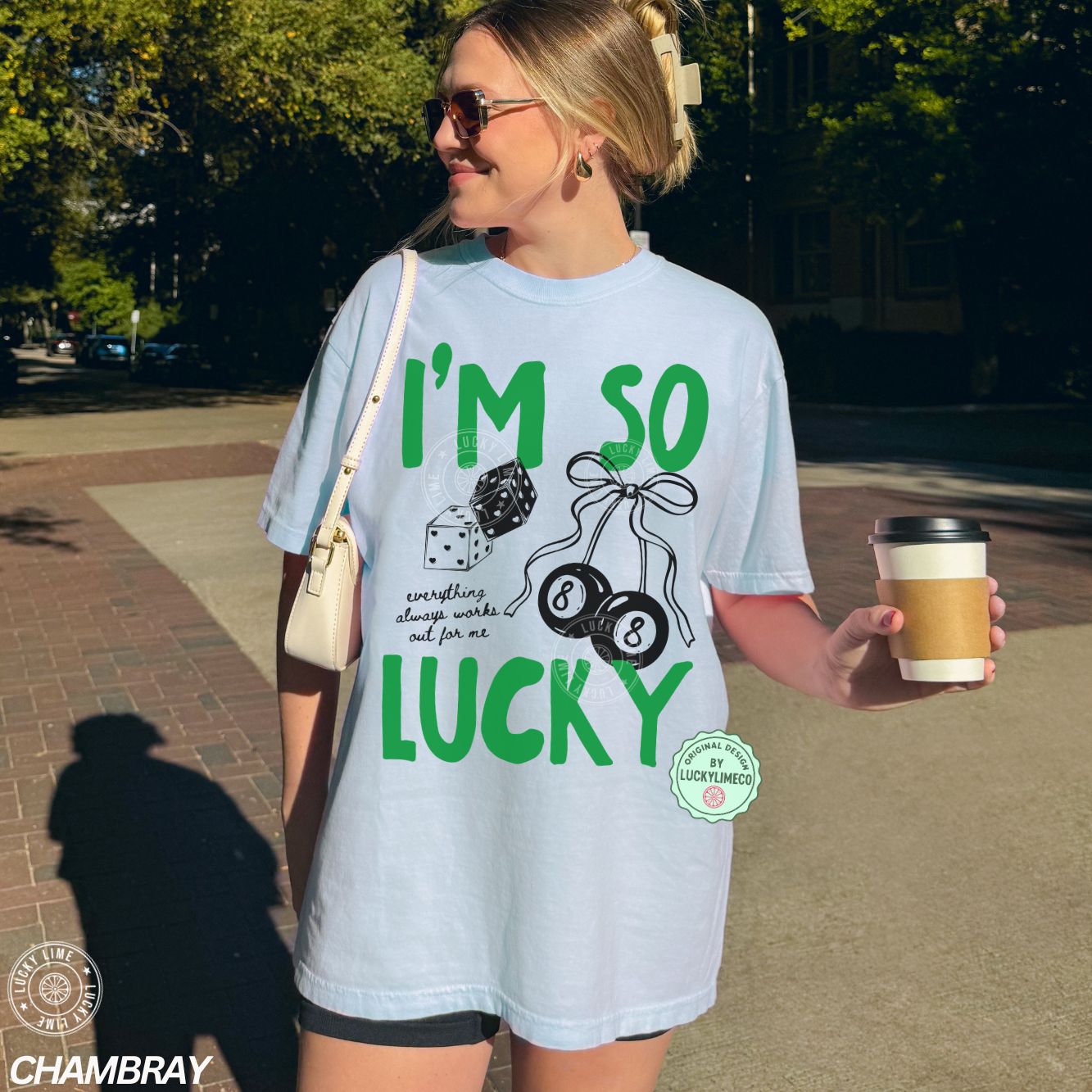 Woman wearing a light blue t-shirt with 'I'm so lucky' text and graphics, holding a coffee cup outdoors.