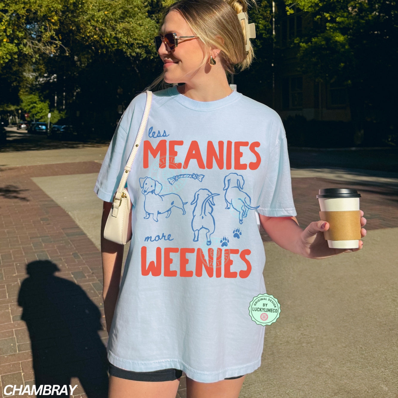 Woman wearing a light blue t-shirt with dachshund  graphics and the phrase ‘less meanies, more weenies’, holding a coffee cup on a sidewalk.