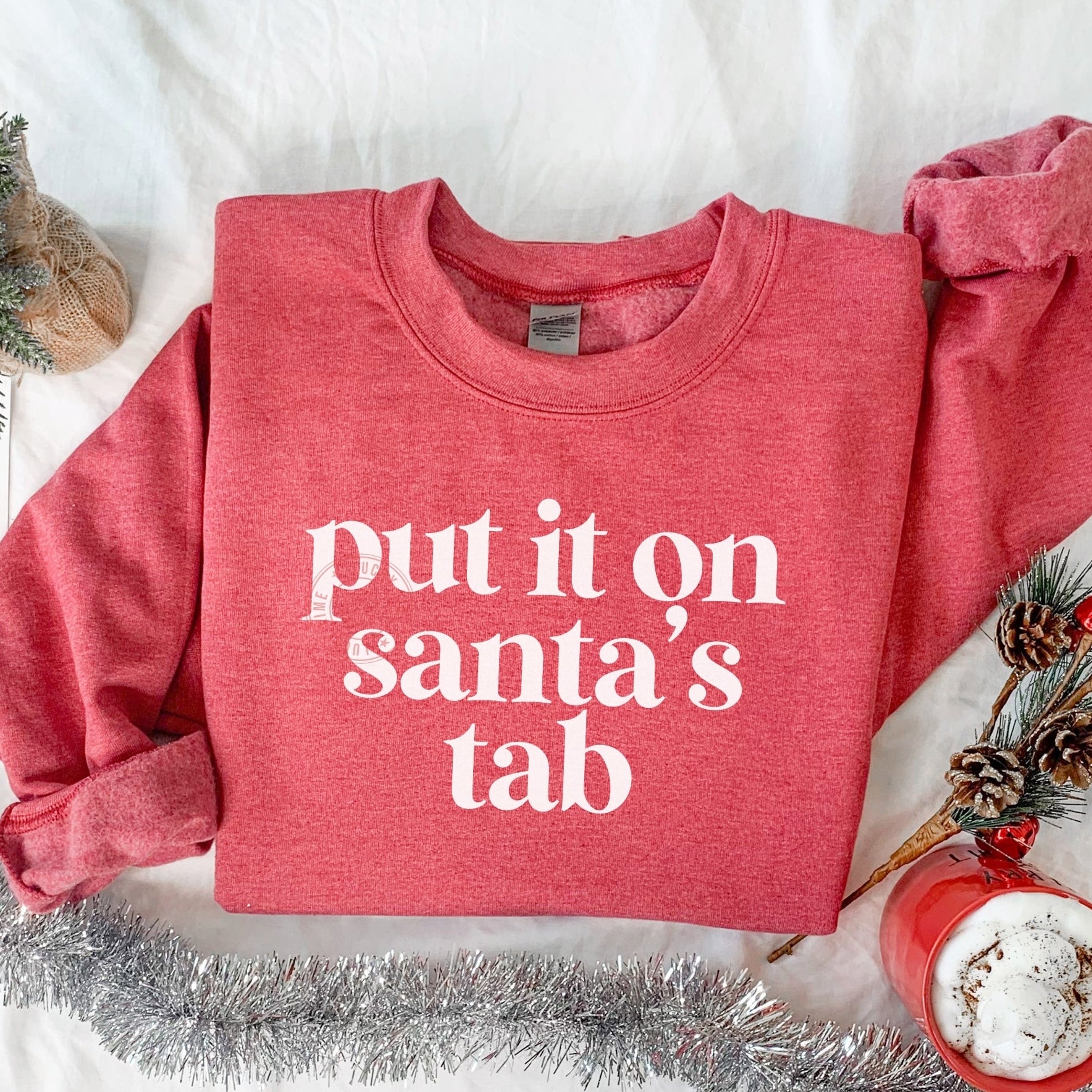 Red sweatshirt with 'put it on Santa's tab' text on a white surface with Christmas decorations.