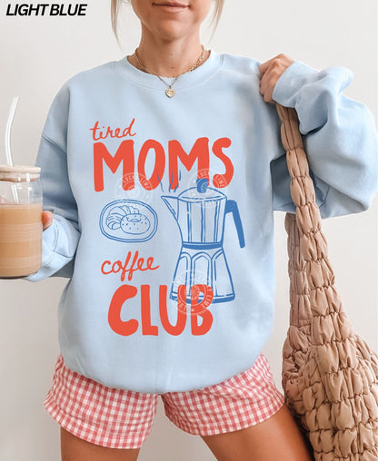 Person wearing a light blue sweatshirt with 'tired moms coffee club' text and red shorts, holding a bag and a drink.