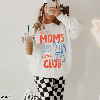 Person wearing a white sweatshirt with 'tired moms coffee club' text, holding a drink.