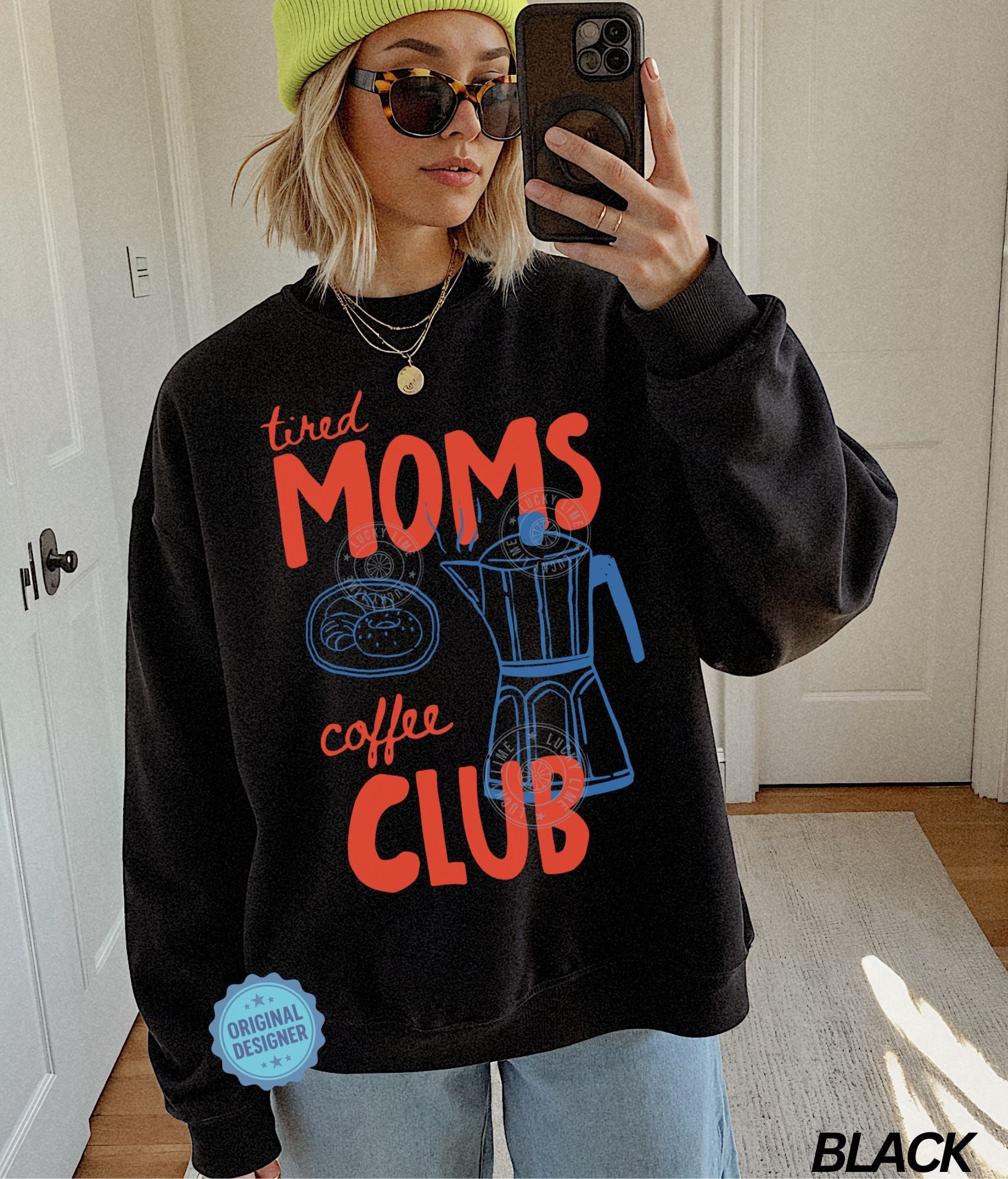 Person wearing a black sweatshirt with 'tired moms coffee club' text and graphics, taking a mirror selfie.
