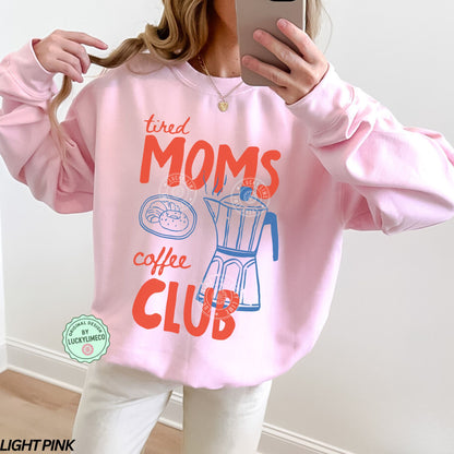 Person wearing a light pink sweatshirt with 'tired moms coffee club' text and graphics.