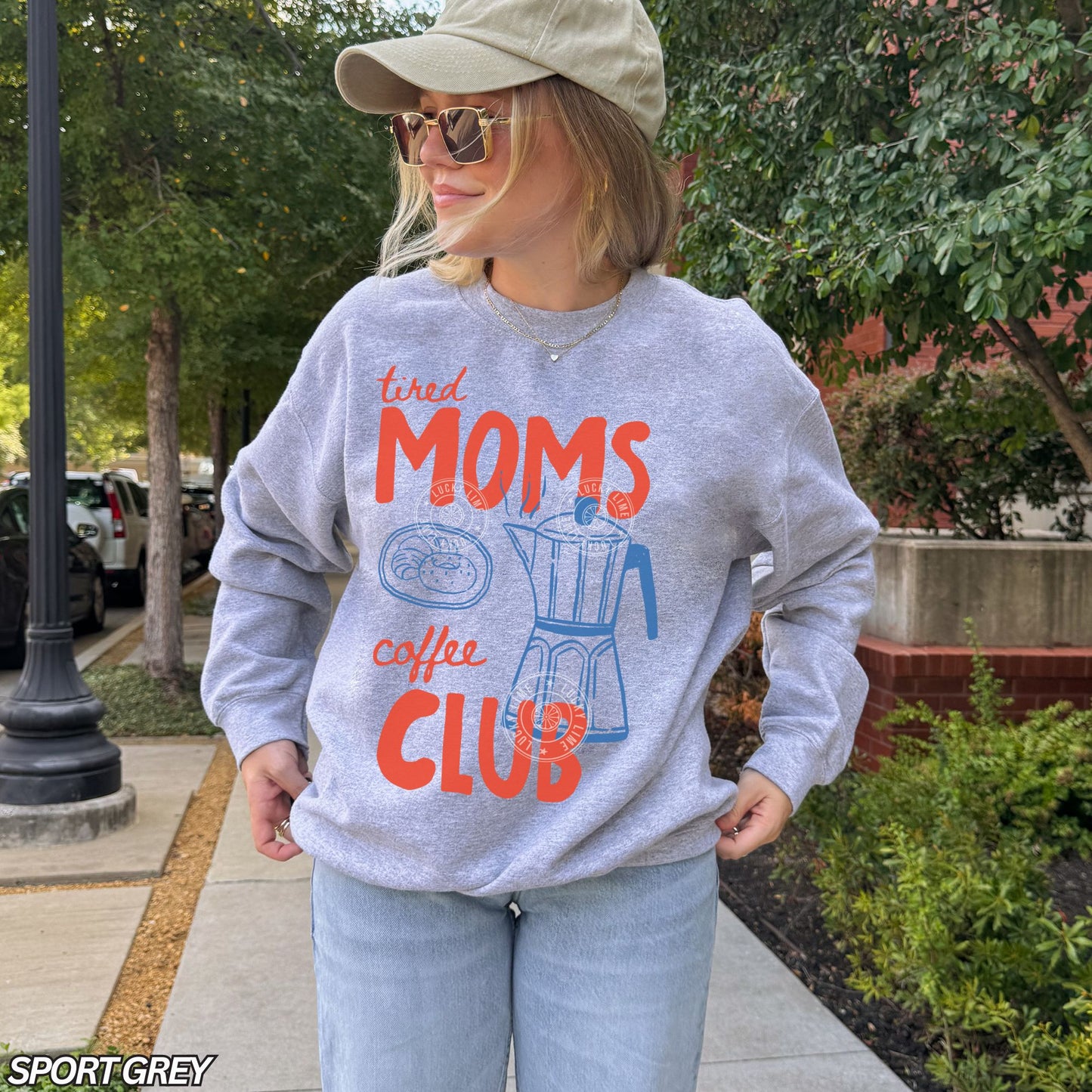 Person wearing a sport grey sweatshirt with 'tired moms coffee club' text outdoors.