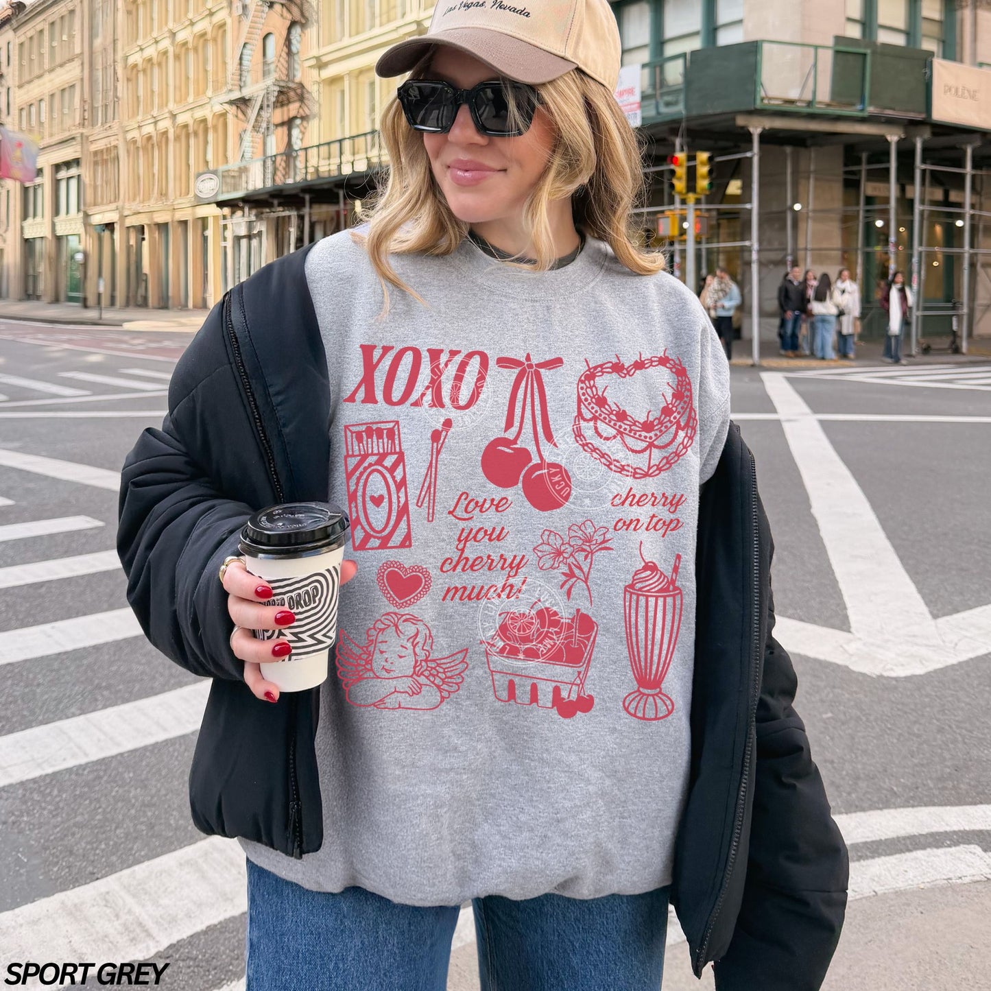 Person wearing a gray sweatshirt with red Valentine graphics and text, holding a coffee cup on a city street.
