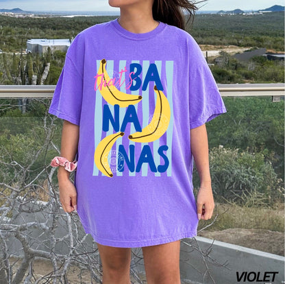 Person wearing a purple t-shirt with banana graphics and text outdoors.