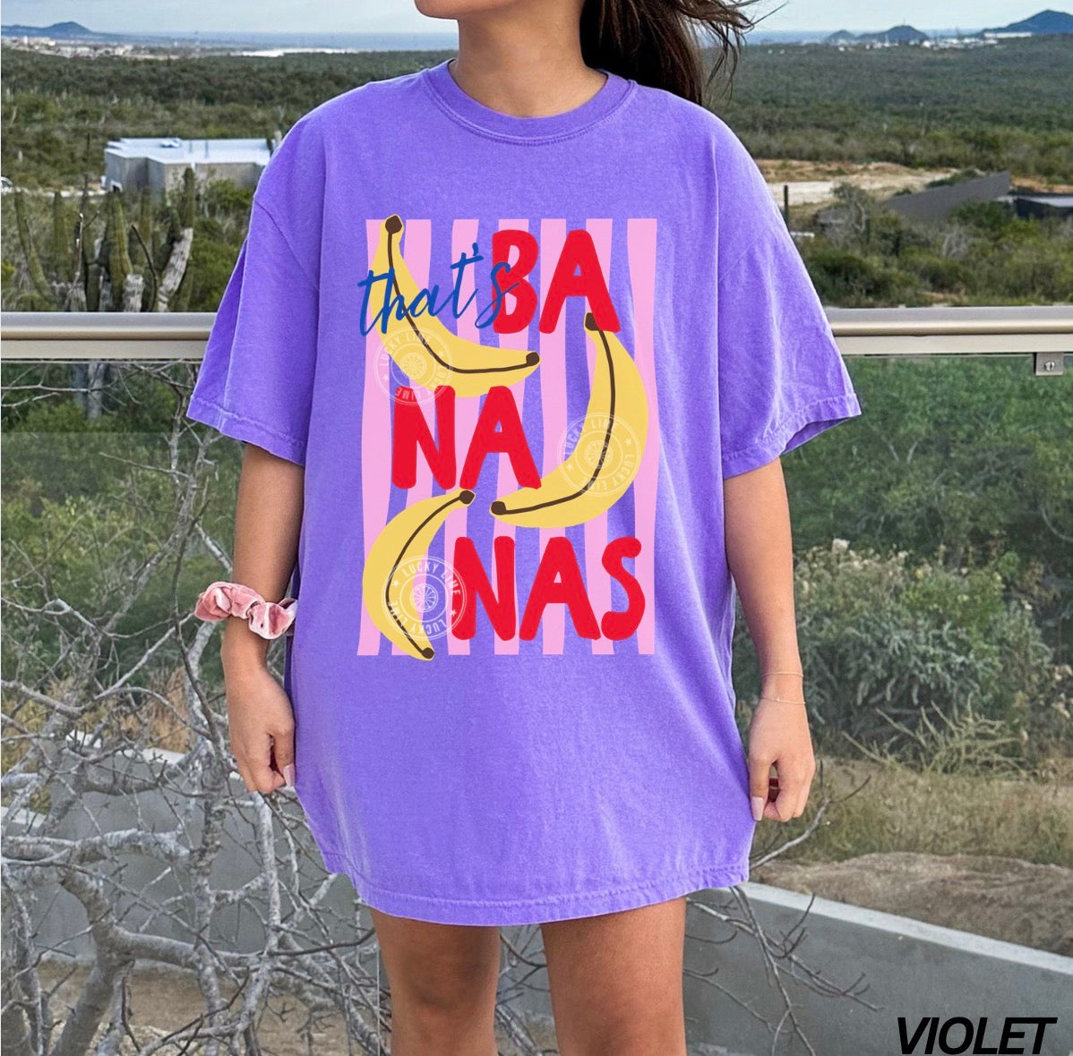 Person wearing a purple t-shirt with colorful banana graphic and text outdoors.