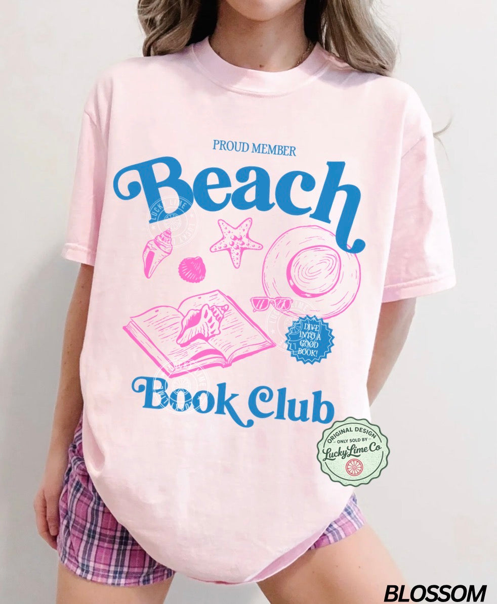 Person wearing a pink t-shirt with 'Beach Book Club' design and plaid shorts on a light gray background.