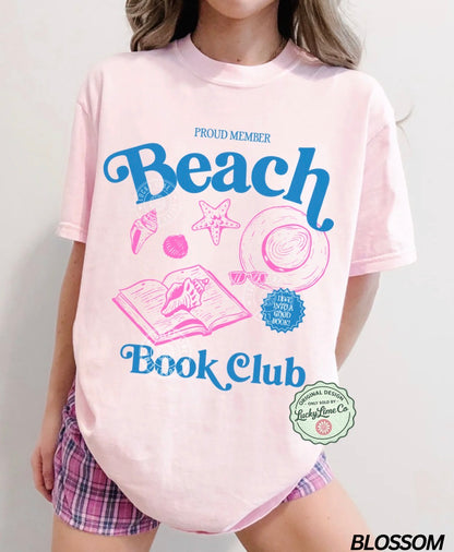 Person wearing a pink t-shirt with 'Beach Book Club' design and plaid shorts on a light gray background.