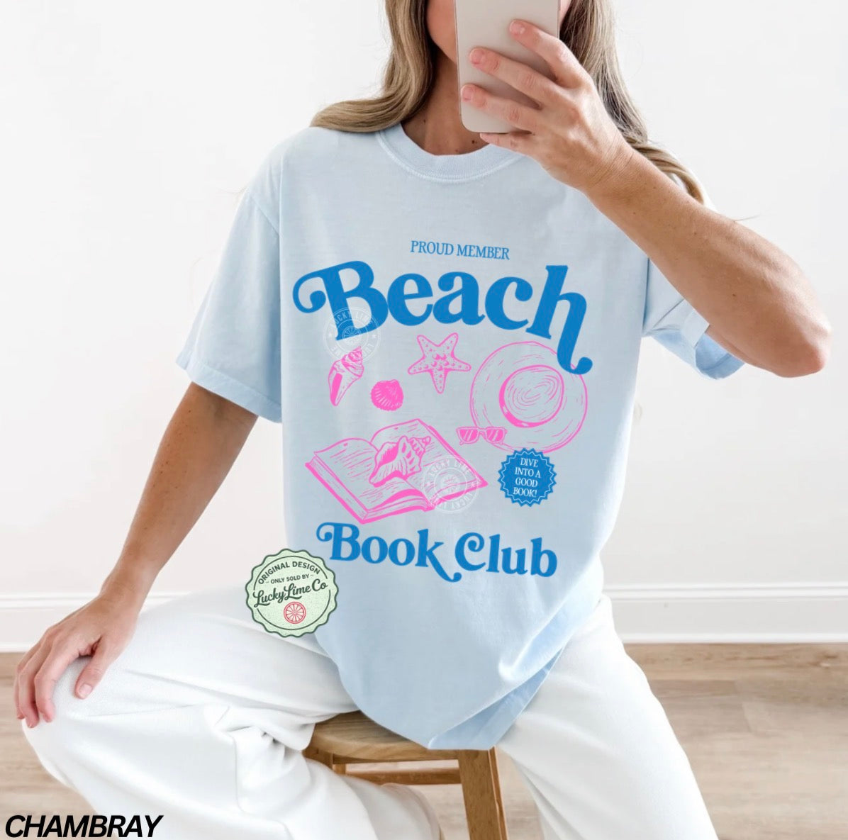 Person wearing a light blue t-shirt with 'Beach Book Club' design, sitting on a wooden stool.