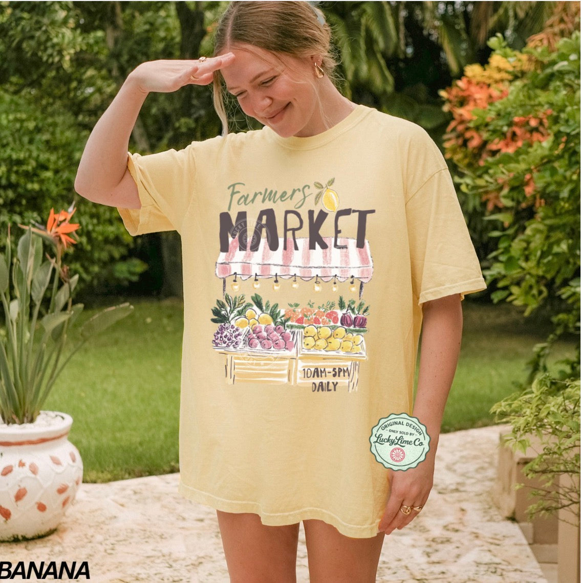Woman wearing a yellow 'Farmers Market' t-shirt outdoors with greenery in the background