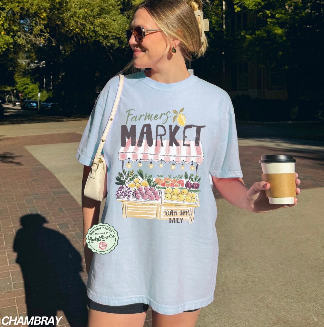Woman wearing a light blue t-shirt with 'Farmers Market' design, holding a coffee cup outdoors.