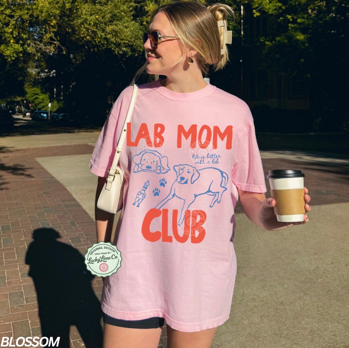 Woman wearing a pink 'Lab Mom Club' t-shirt holding a coffee cup outdoors.