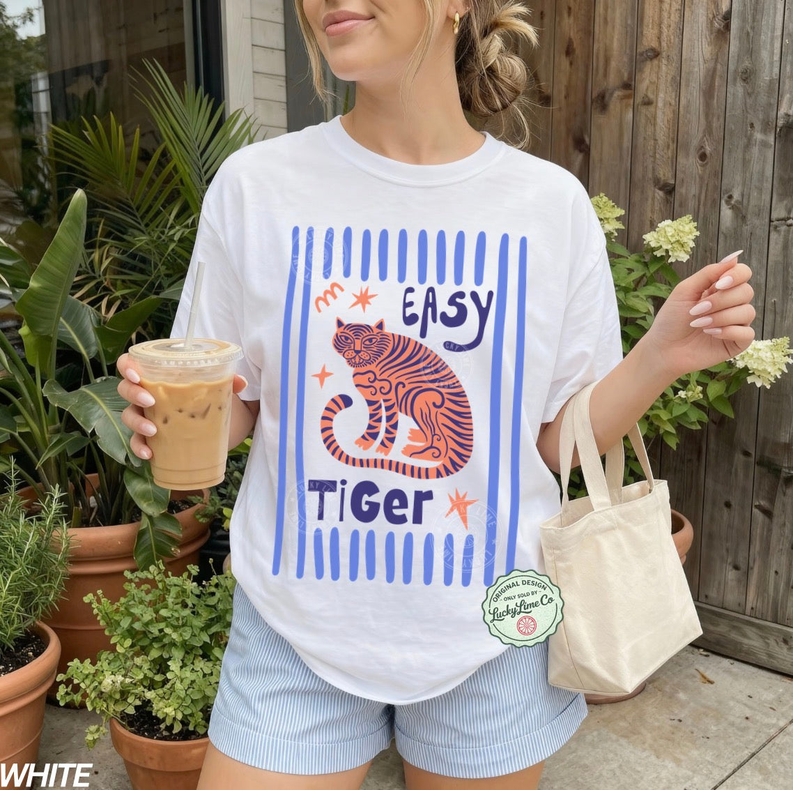 Person wearing a white t-shirt with a tiger graphic and 'Easy Tiger' text, holding a drink and a bag outdoors.