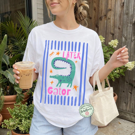 Person wearing a white t-shirt with an alligator graphic and text saying “later gator”, holding a drink and a bag outdoors.