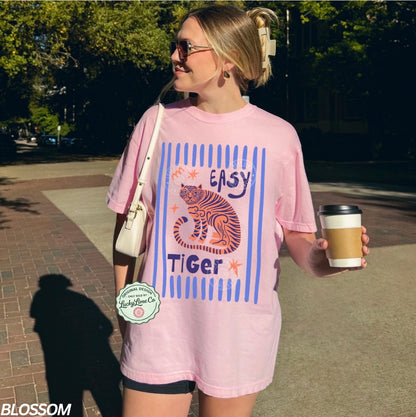 Woman wearing a pink t-shirt with a tiger graphic and text, holding a coffee cup outdoors.