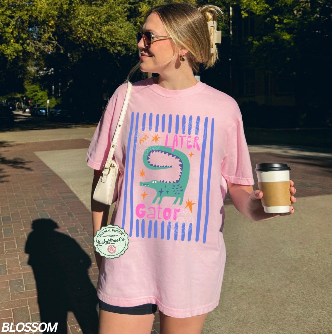Person wearing a pink t-shirt with an alligator graphic and text saying “later gator”, holding a drink and a bag outdoors.