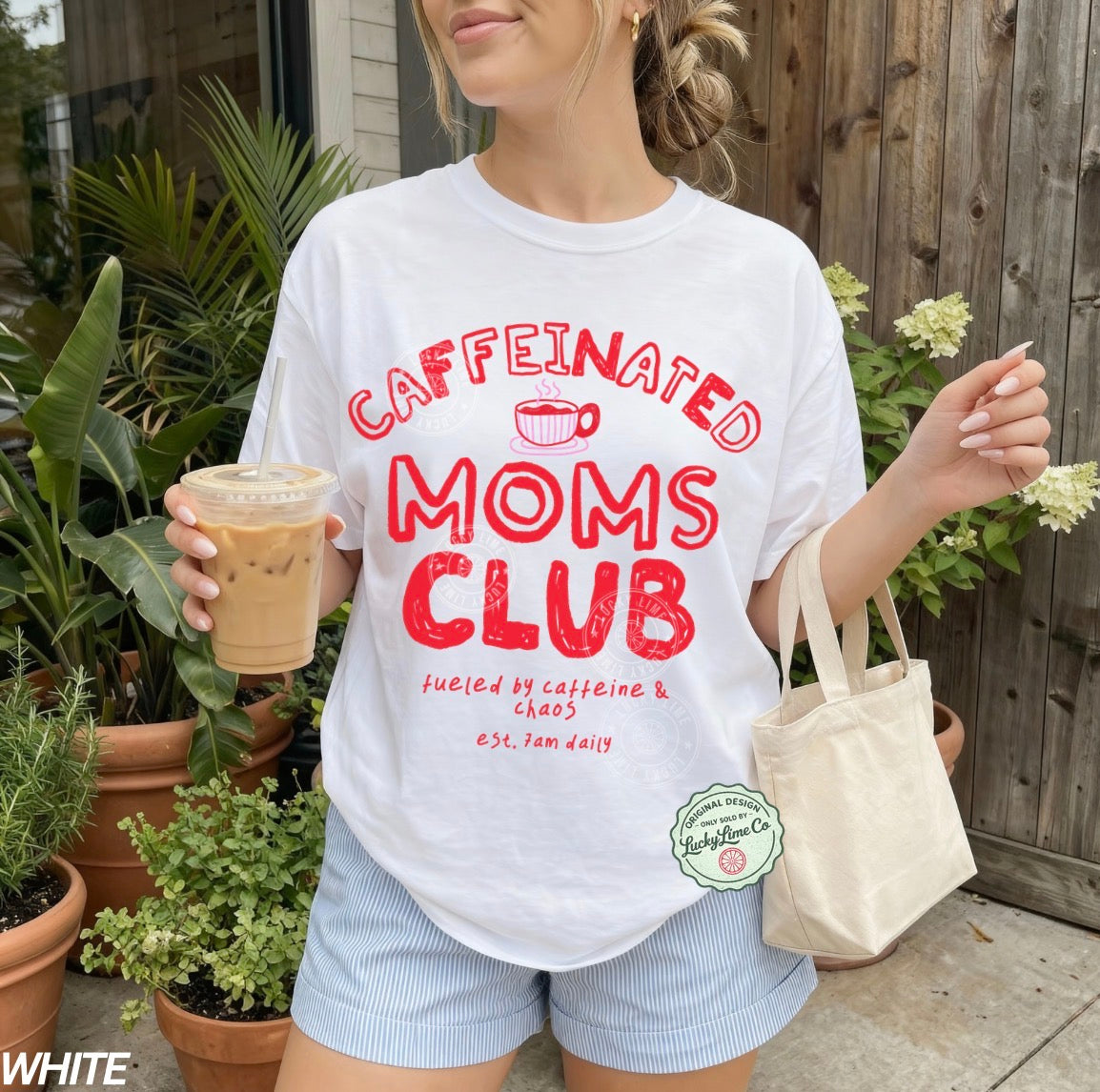 Person wearing a 'Caffeinated Moms Club' t-shirt holding a drink and a tote bag outdoors.