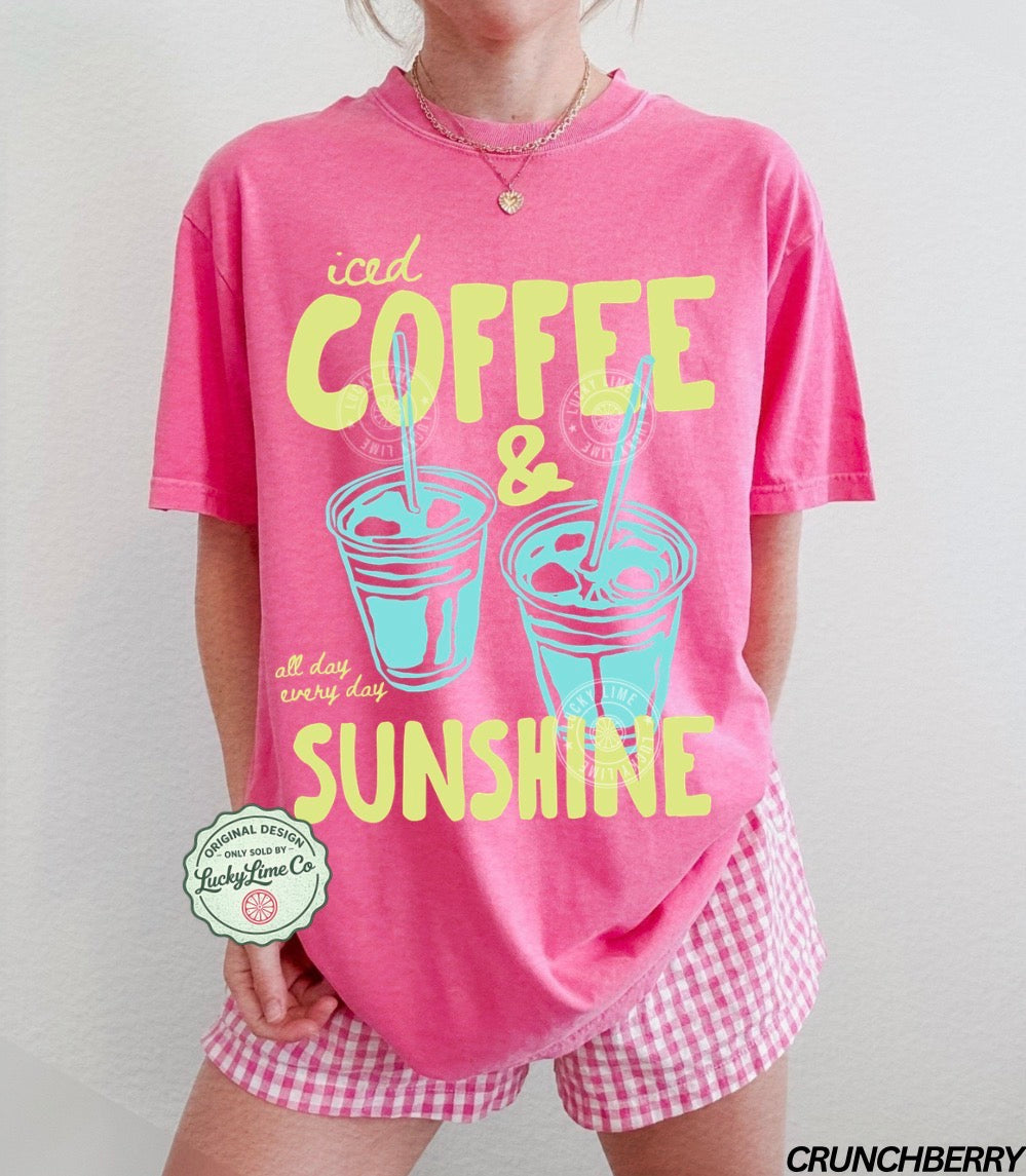 Person wearing a pink t-shirt with 'iced coffee & sunshine' text and checkered shorts on a plain background.
