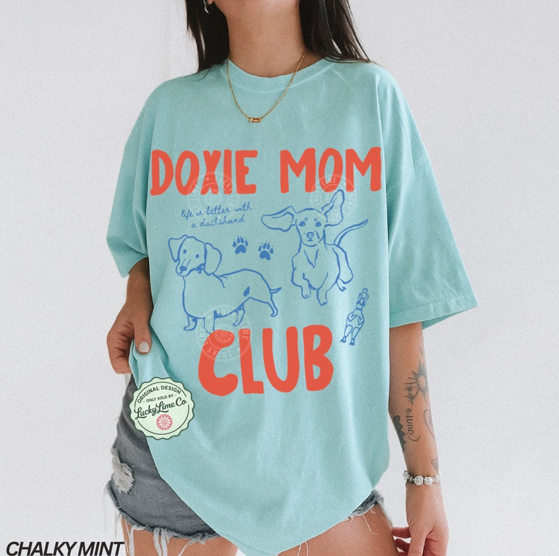 Person wearing a light blue t-shirt with 'Doxie Mom Club' text and dachshund illustrations on a white background.