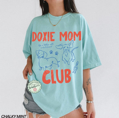 Person wearing a light blue t-shirt with 'Doxie Mom Club' text and dachshund illustrations on a white background.