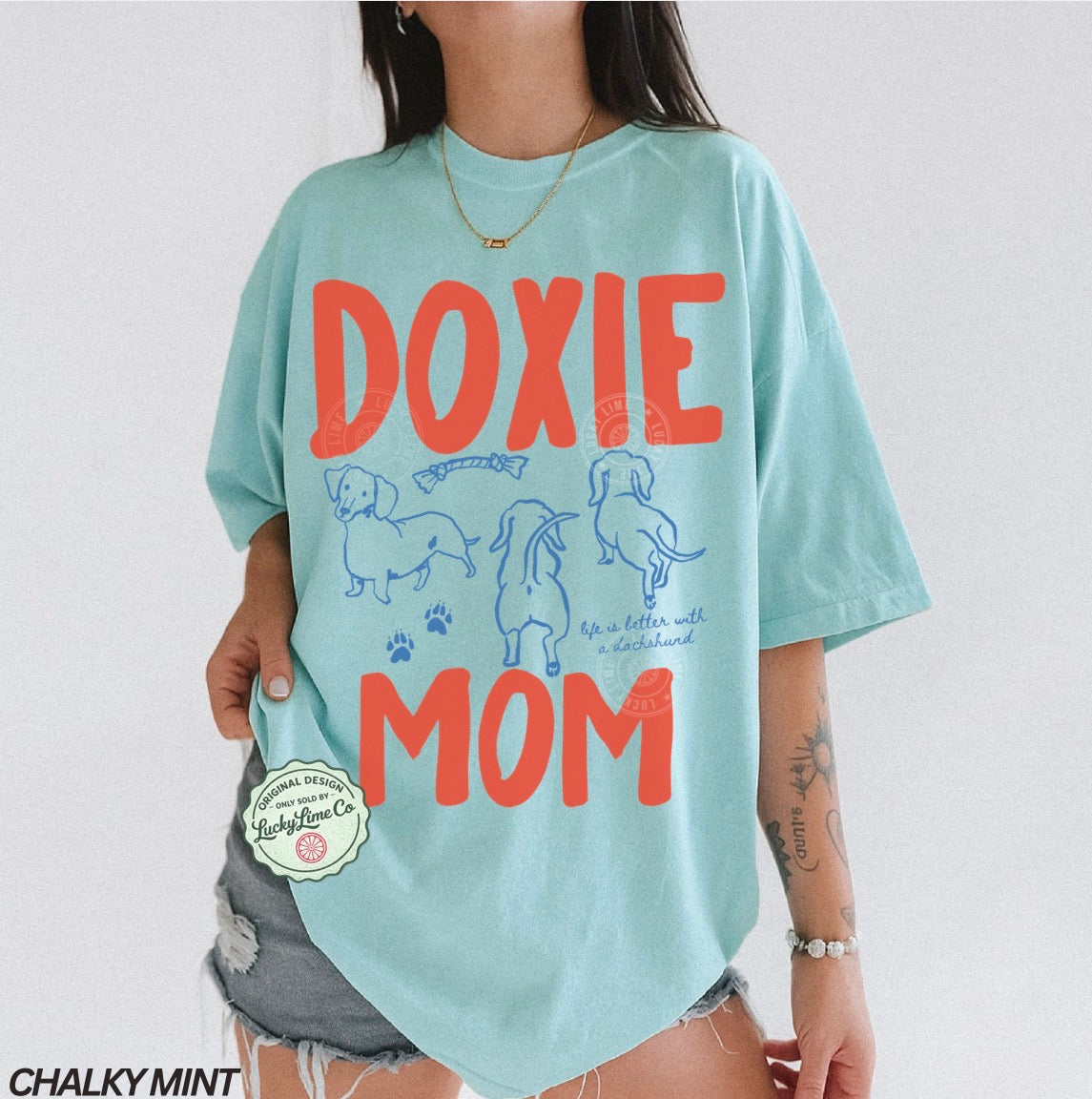 Person wearing a light blue t-shirt with 'Doxie Mom' text and dachshund illustrations on a white background.