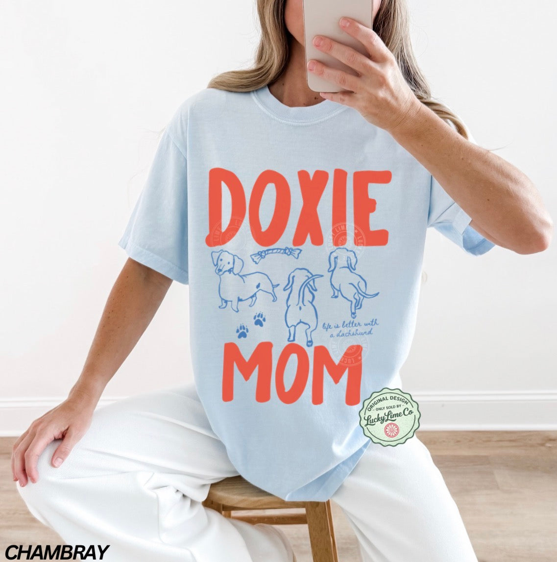 Person wearing a light blue t-shirt with 'Doxie Mom' text and dog illustrations, sitting on a wooden stool.