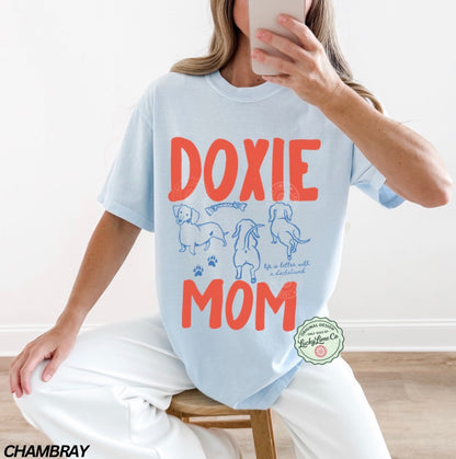 Person wearing a light blue t-shirt with 'Doxie Mom' text and dog illustrations, sitting on a wooden stool.