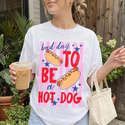 Person wearing a white t-shirt with a colorful graphic and text, holding a drink and a bag outdoors.