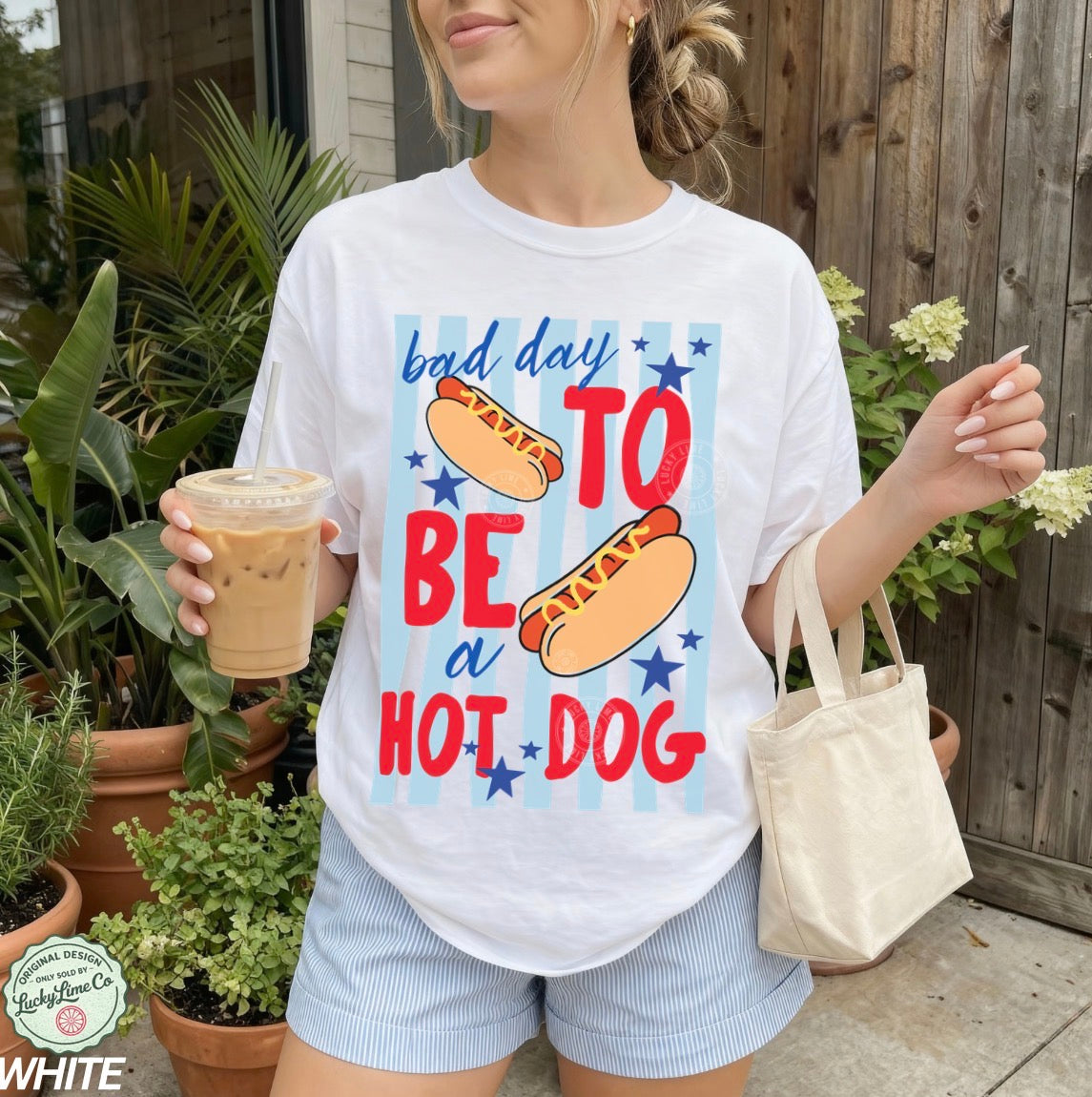 Person wearing a white t-shirt with a colorful graphic and text, holding a drink and a bag outdoors.