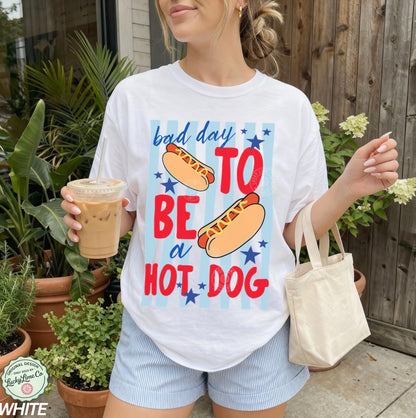 Person wearing a white t-shirt with a colorful graphic and text, holding a drink and a bag outdoors.