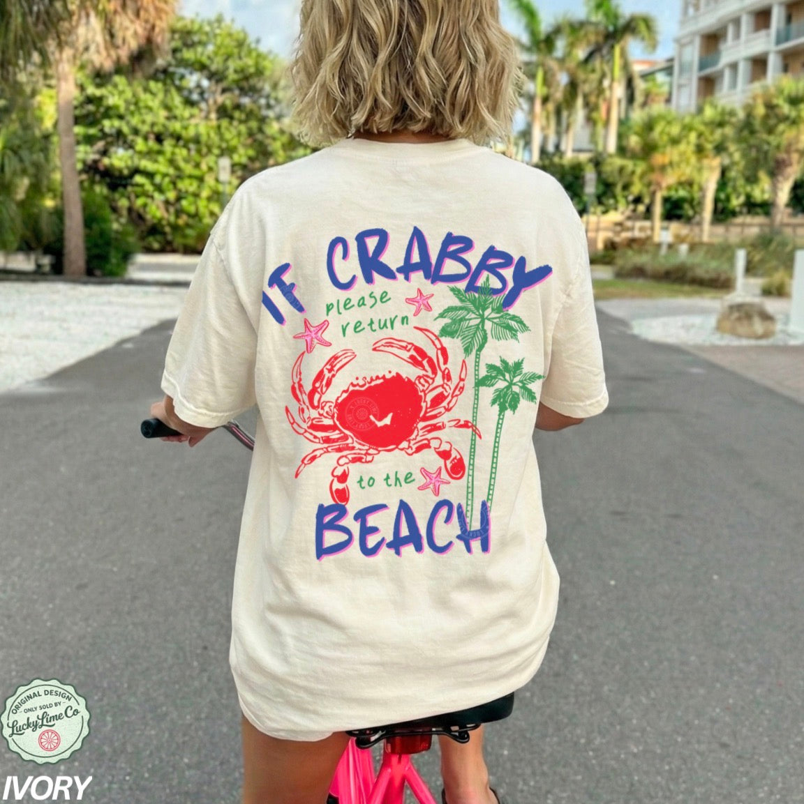 Person wearing a t-shirt with a beach-themed design and text, standing on a path with greenery and a building in the background.